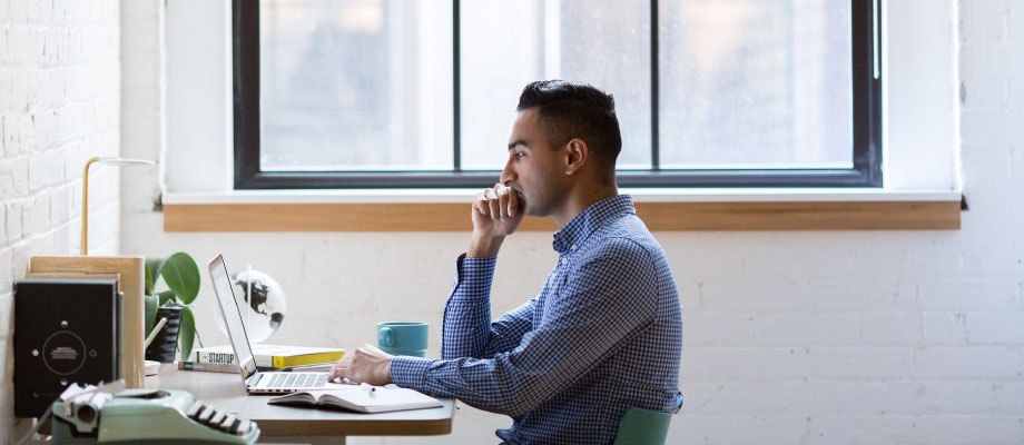 guy wearing blue button up sitting at desk, looking at laptop, window behind him