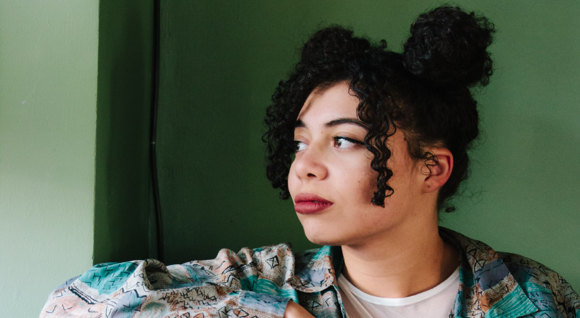 Dark-haired woman with ringlets pulled into top bun stares out a window by a green wall wondering about fuzzy teeth