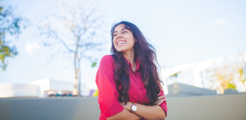 Brunette woman wearing a red blouse folds her arms and smiles as she looks to the side in a parking lot