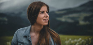 Brunette woman wearing a denim blouse smiles while looking off to the side in front of a mountain on a cloudy day