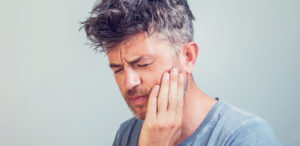 Middle-aged man wearing a gray shirt touches his cheek with his eyes closed due to sensitivity and pain from a lost filling