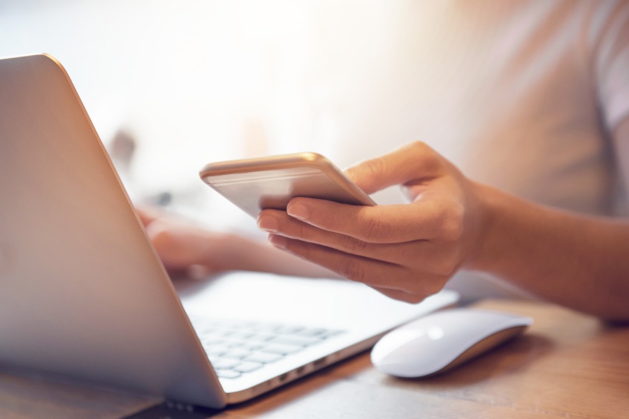 Closeup of a person's hand holding their cell phone while on their laptop to stay informed about the coronavirus